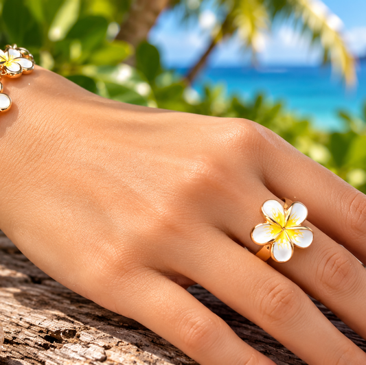 A close-up of a person's hand wearing a gold bracelet and matching flower ring, with tropical beach scenery and white plumeria flowers in the background.