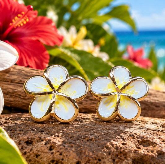 A pair of gold-plated flower stud earrings with white enamel petals and yellow centers, resting on a brown rock with tropical flowers and a beach background.