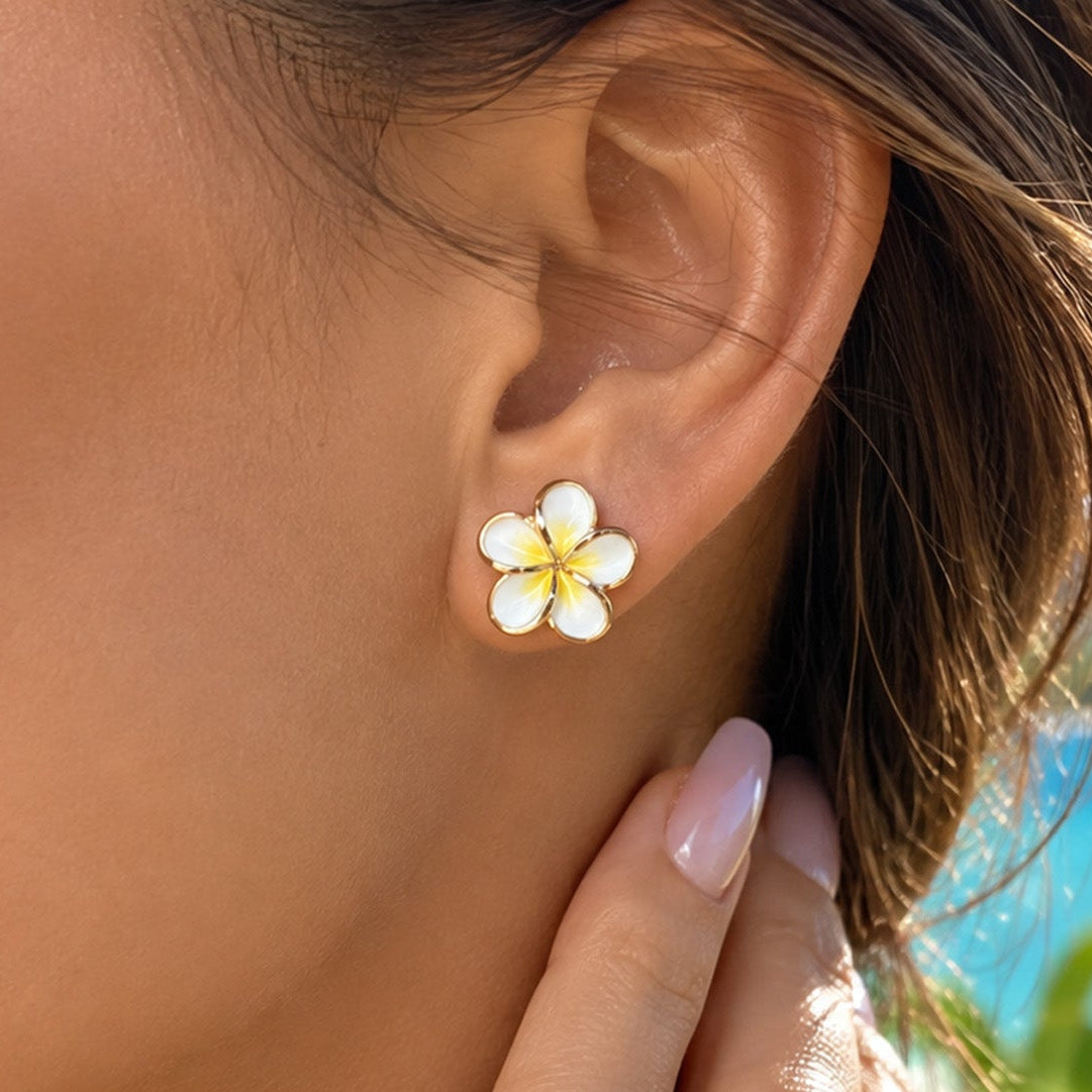 A close-up of a woman's ear wearing a small white flower stud earring with yellow center details, set against a tropical beach background with blue water and green foliage.