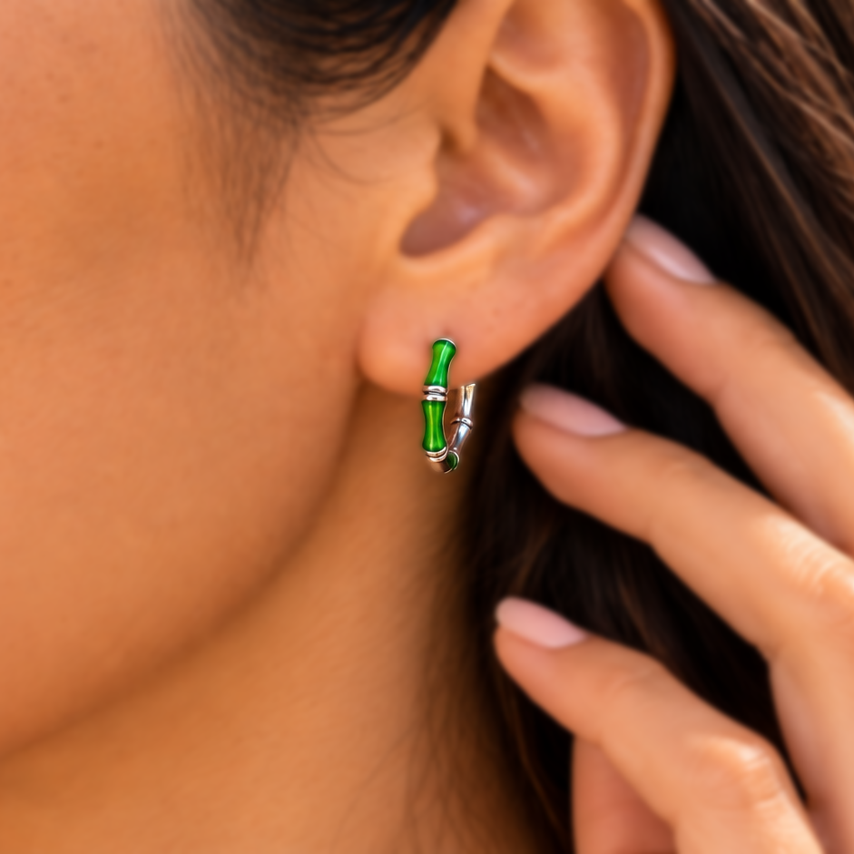 A close-up lifestyle shot of a woman wearing a small silver hoop earring with a bright green bamboo leaf accent, set against a blurred tropical beach background.