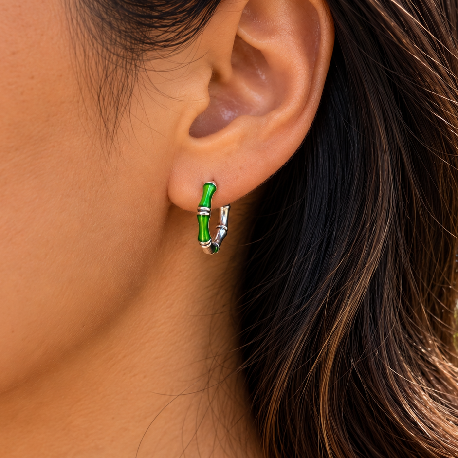 A close-up of a woman's ear wearing a small hoop earring with a bright green center and silver-toned outer edges, set against a blurred tropical beach background.