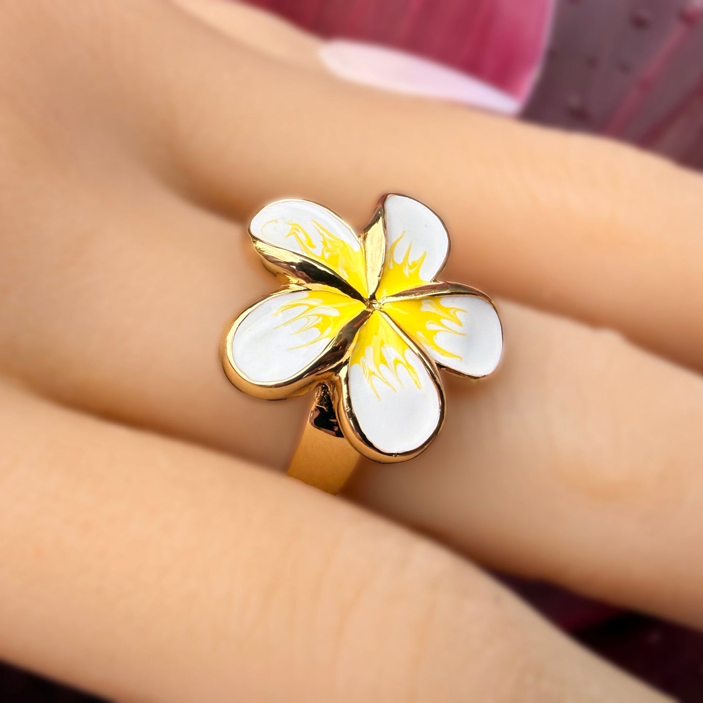 A gold-plated ring featuring a white and yellow plumeria flower design, shown worn on a person's finger.