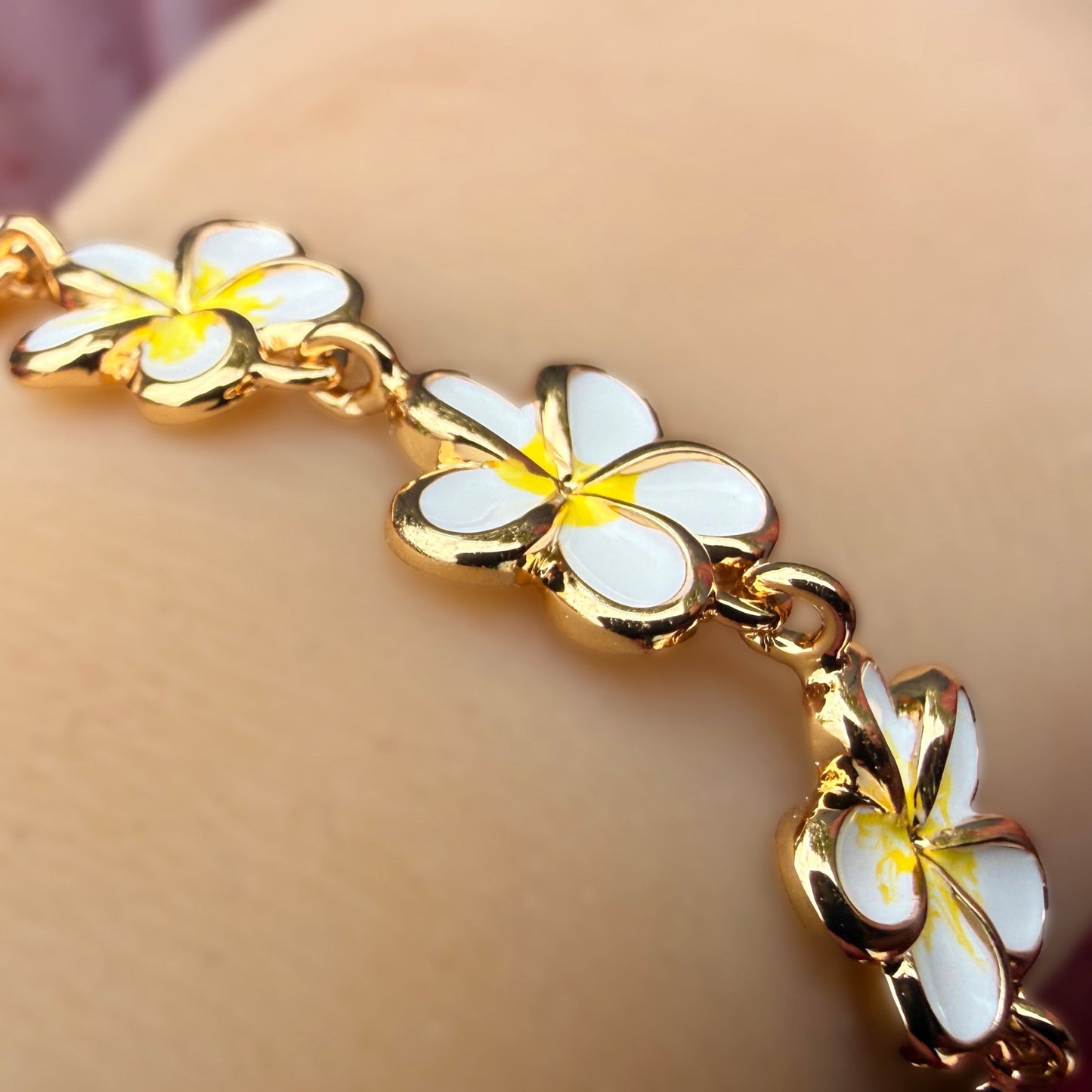 A gold-plated bracelet featuring white and yellow plumeria flower charms, shown worn on a person's wrist.