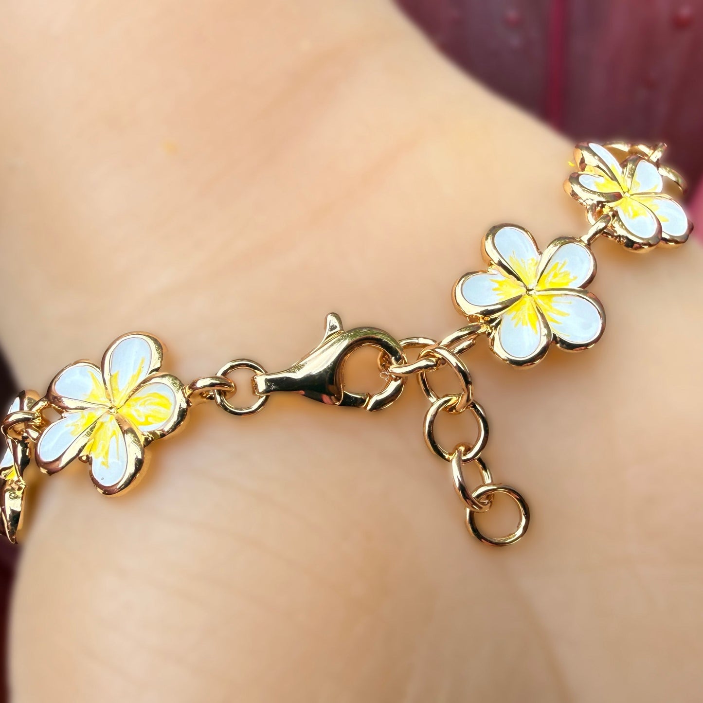 A gold-plated chain bracelet with four white and yellow plumeria flower charms, shown worn on a person's wrist.