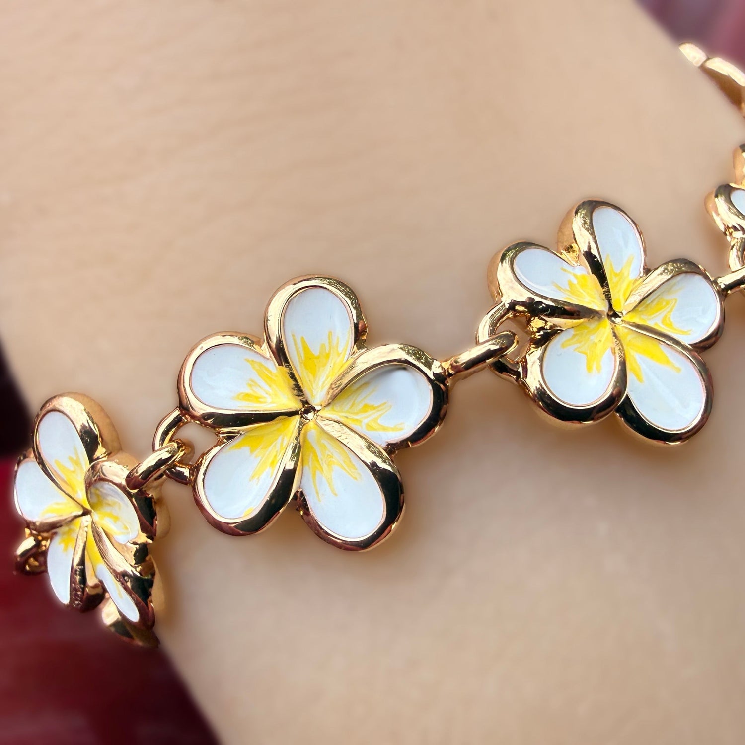 A gold-plated bracelet featuring five white enamel plumeria flower charms with yellow centers, shown worn on a wrist.