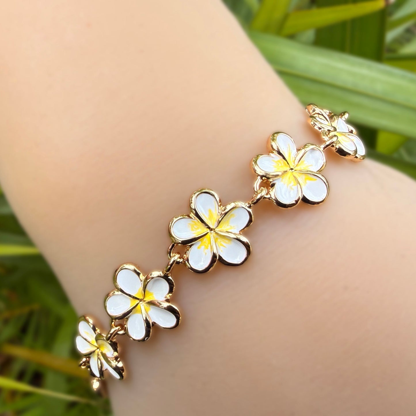 A gold-plated bracelet featuring five white plumeria flower charms with yellow centers, worn on a person's wrist against a background of green tropical leaves.