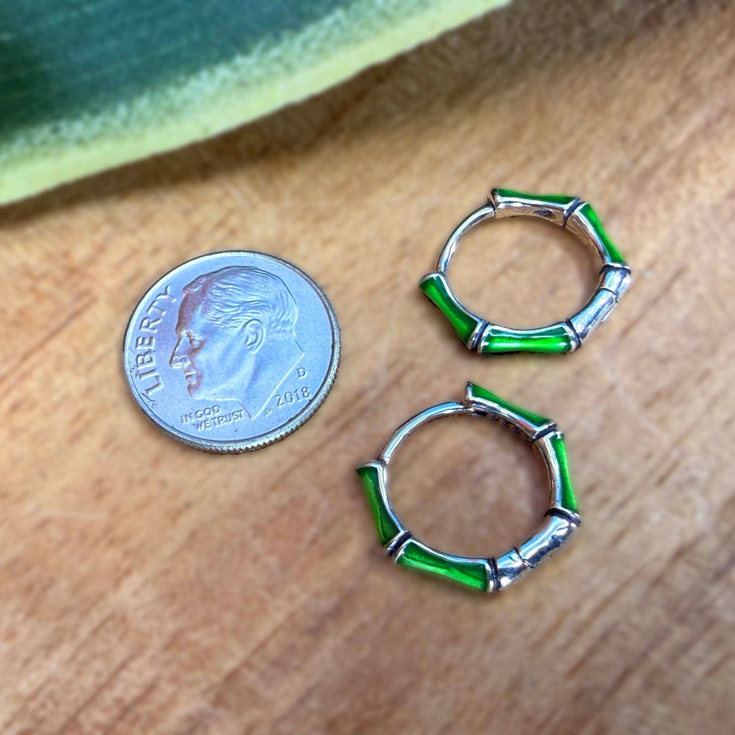 A pair of small hoop earrings with green bamboo-style segments and silver metal accents, shown on a wooden surface next to a U.S.