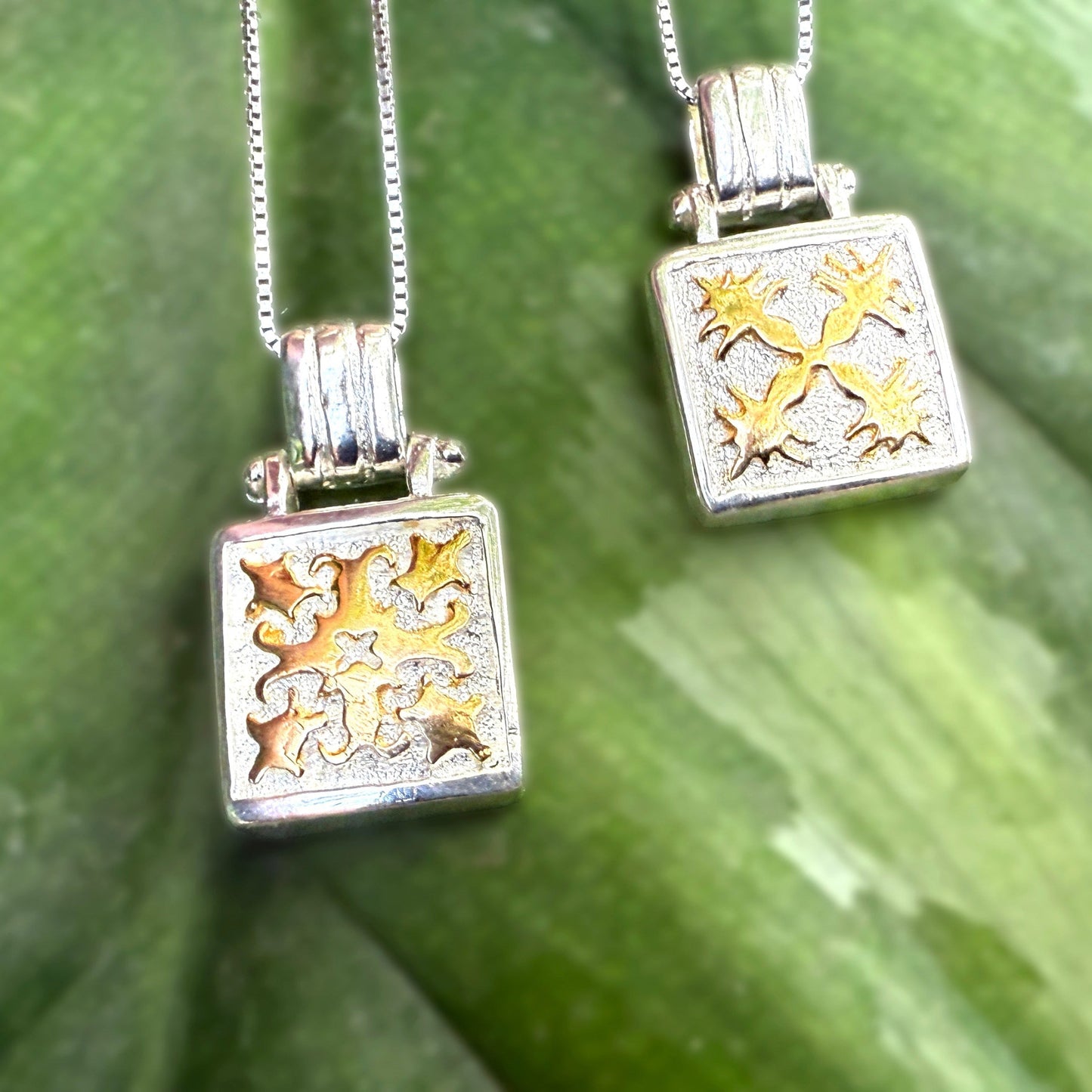Two square pendants with gold and silver designs on a green leaf background