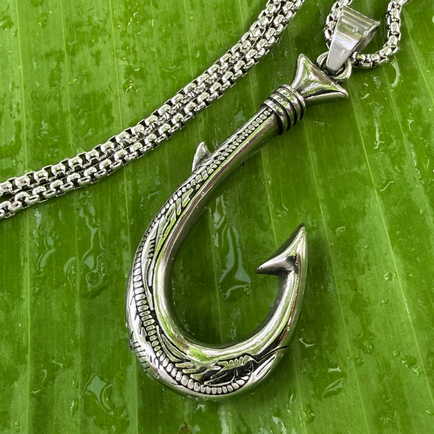 A silver-toned fishhook pendant necklace with a textured, carved design, resting on a glossy green banana leaf with water droplets.