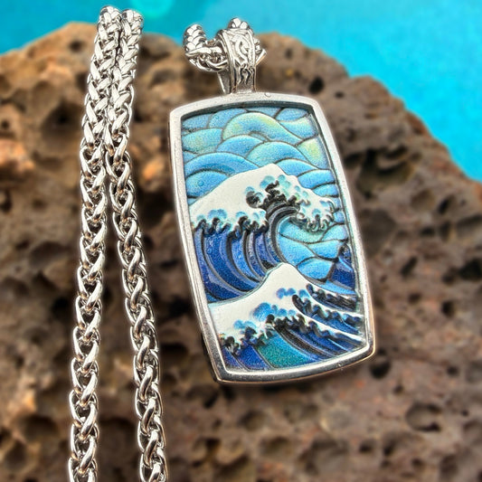 A silver-toned rectangular pendant necklace featuring an enamel-style depiction of the Great Wave off Kanagawa, displayed on a textured brown rock against a bright turquoise background.