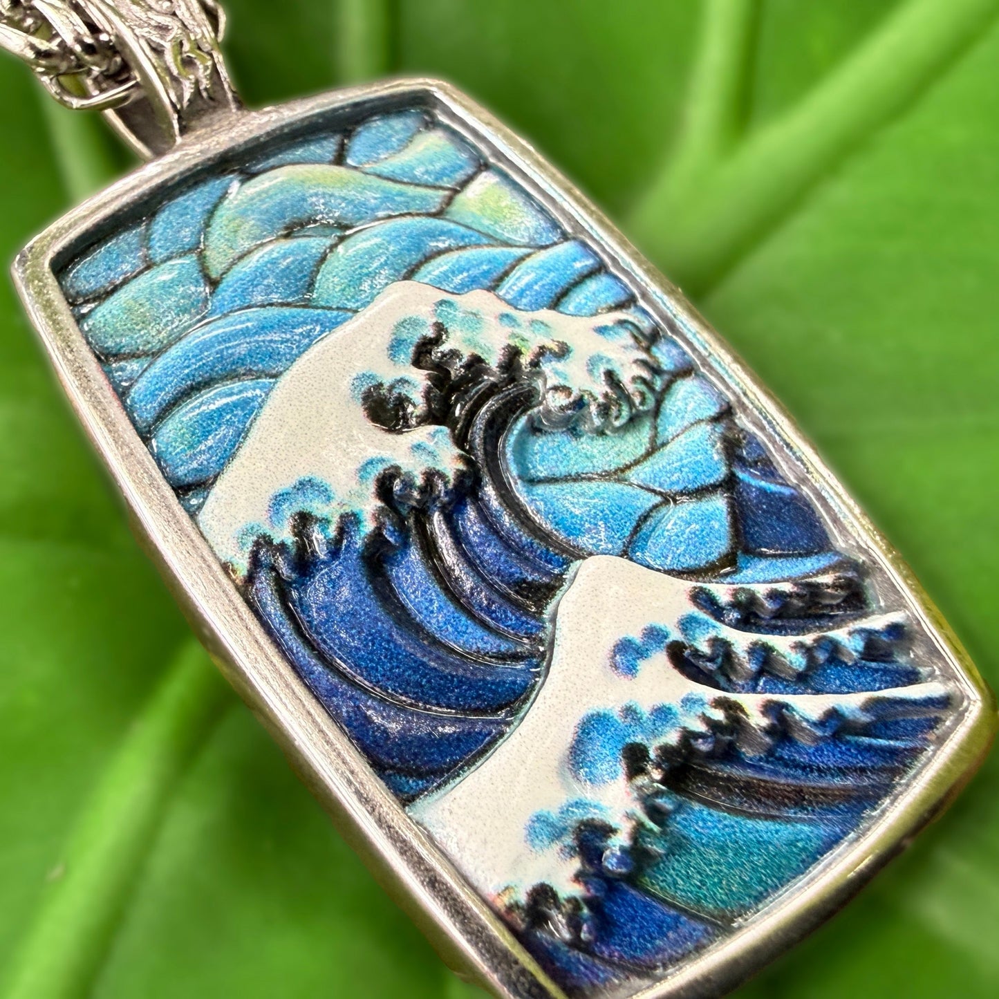 A rectangular pendant featuring a stylized blue and white ocean wave design, photographed close-up against a bright green leaf background.