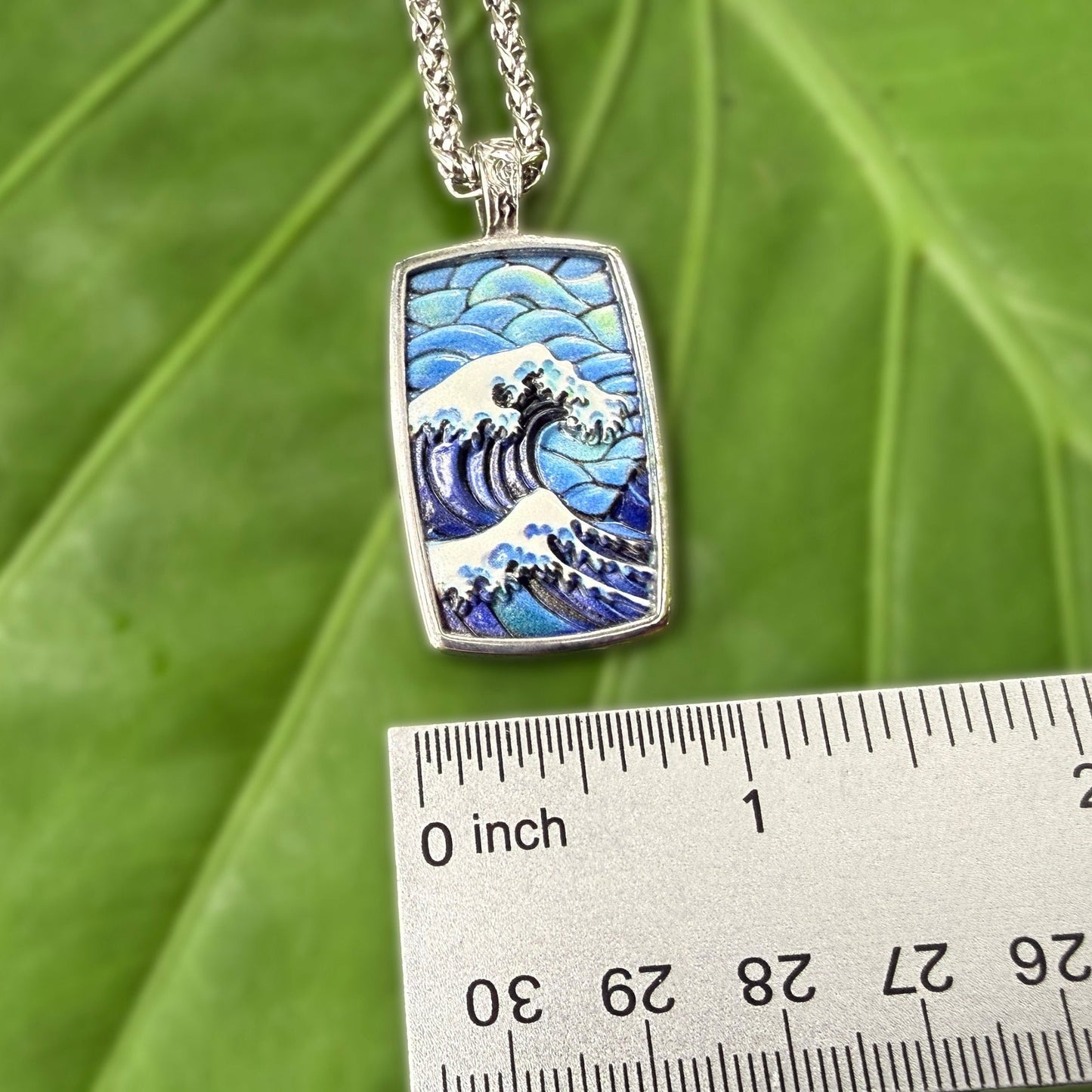 A rectangular silver pendant necklace featuring a blue and white wave design inspired by 'The Great Wave off Kanagawa', displayed on a large green leaf next to a metal ruler for scale.