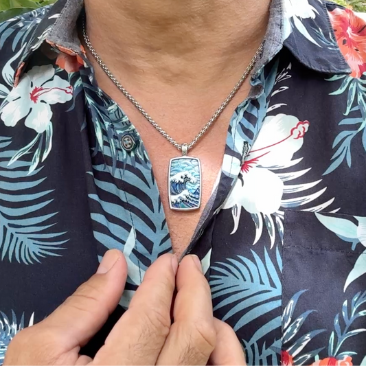 A close-up of a man wearing a silver-toned chain necklace with a rectangular pendant featuring a blue and white wave design. He is wearing a dark navy tropical floral button-down shirt with red and white hibiscus flowers and green leaves, with outdoor foliage visible in the background.