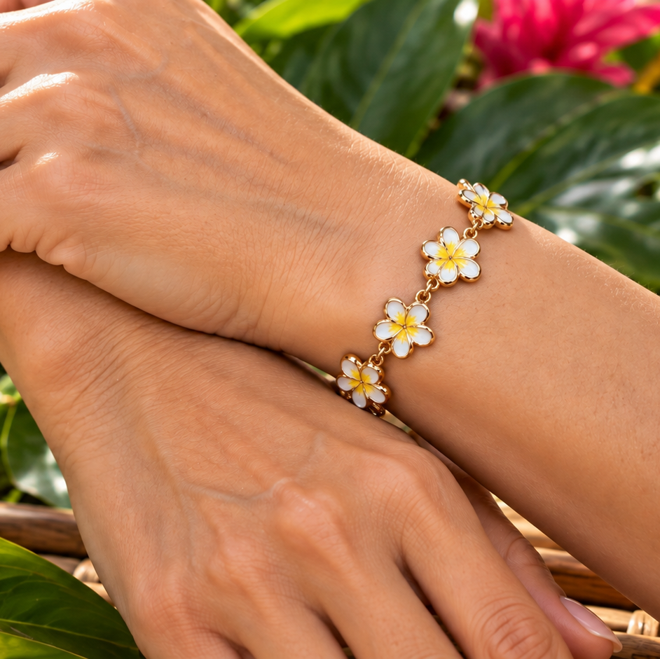 A close-up lifestyle shot of a gold bracelet adorned with white and yellow plumeria flowers worn on a person's wrist, set against a tropical background of green leaves and pink flowers.