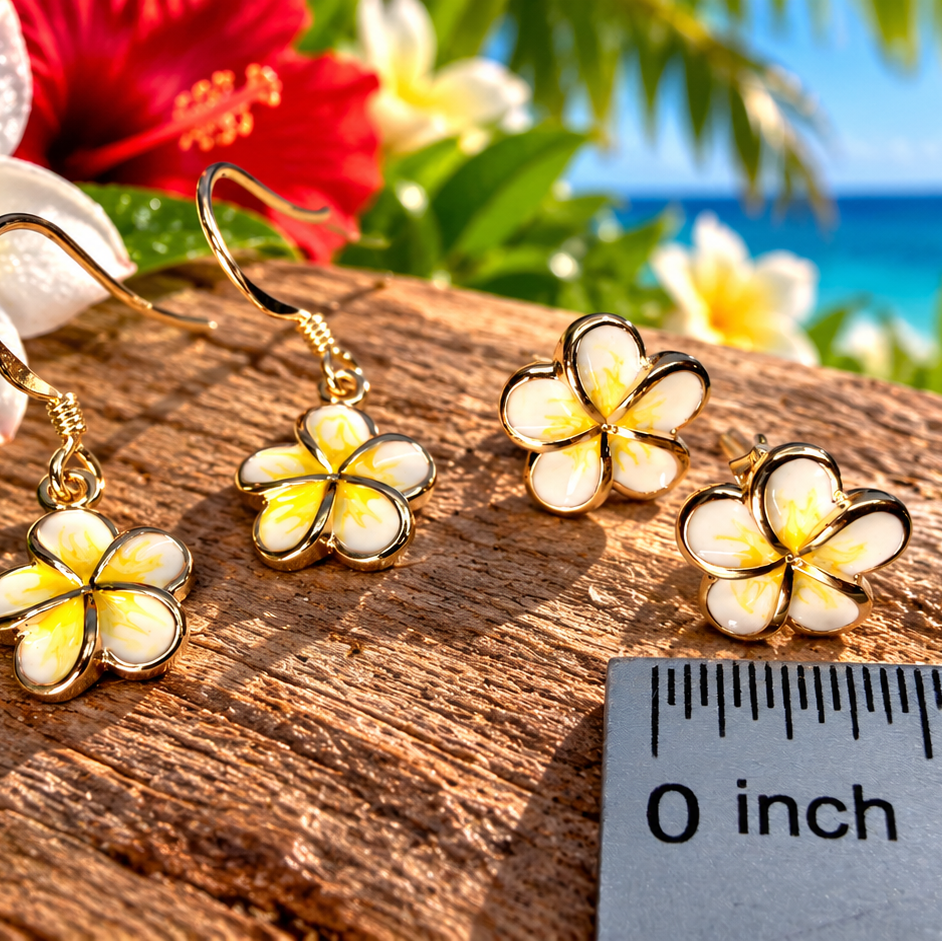 Three gold-plated plumeria flower drop earrings with white enamel centers are displayed on a wooden surface next to a ruler for scale, with tropical flowers and a beach background.