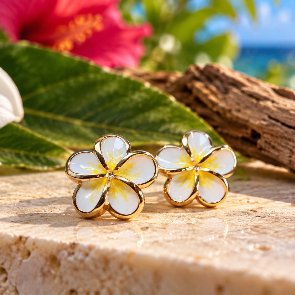 A pair of gold flower stud earrings with white and yellow plumeria-inspired petals, resting on a stone surface with tropical flowers, driftwood, and a blurred ocean background.