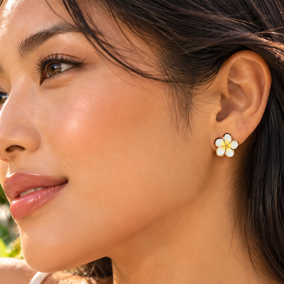 A close-up lifestyle shot of a woman wearing a small white flower stud earring with yellow accents, set against a tropical beach background with palm trees and blue ocean.