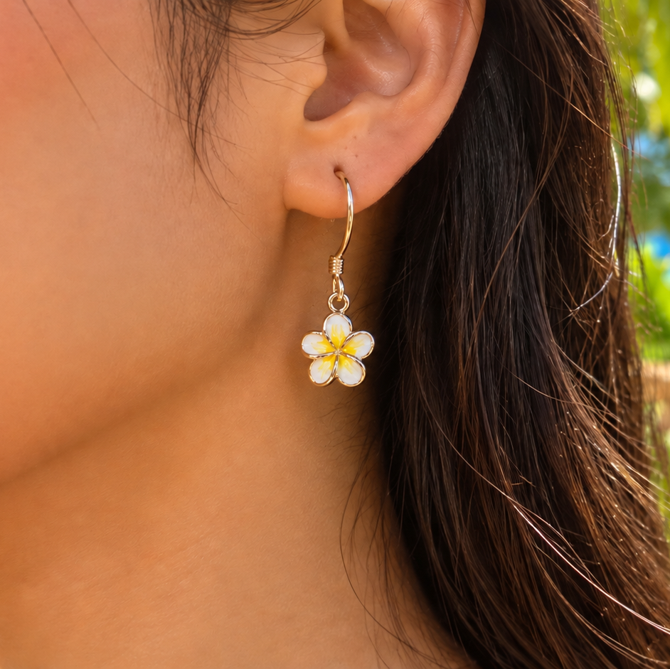 A close-up of a woman's ear wearing a gold hoop earring with a dangling white plumeria flower charm with a yellow center, set against a tropical beach background with palm leaves and ocean.