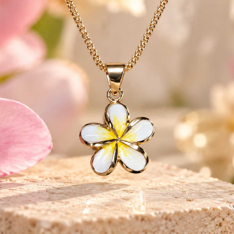 A gold-tone flower pendant necklace with white and yellow enamel details, resting on a stone block surrounded by pink plumeria flowers and a glass shell.