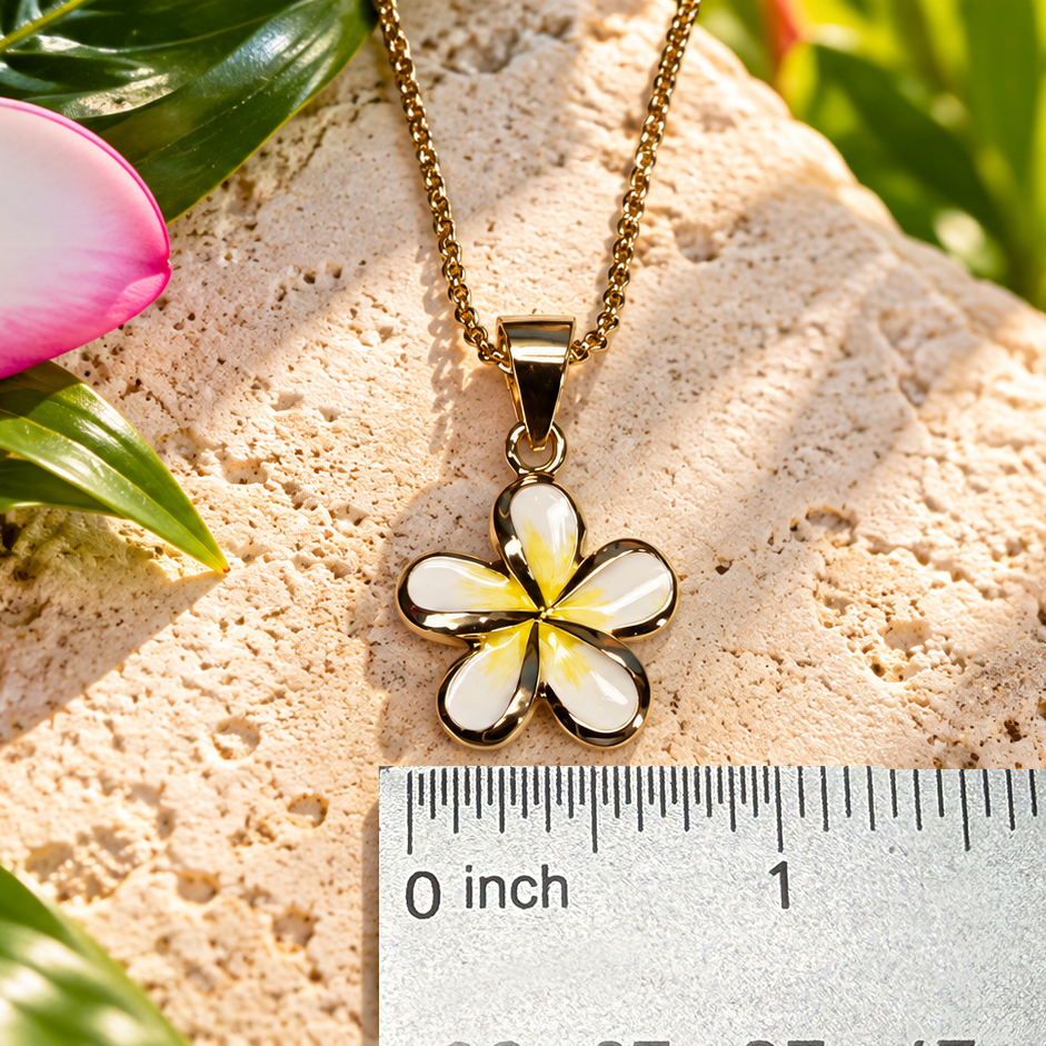 A gold-tone flower pendant necklace with white and yellow enamel details, displayed on a textured stone surface next to a ruler for scale, with tropical flowers in the background.