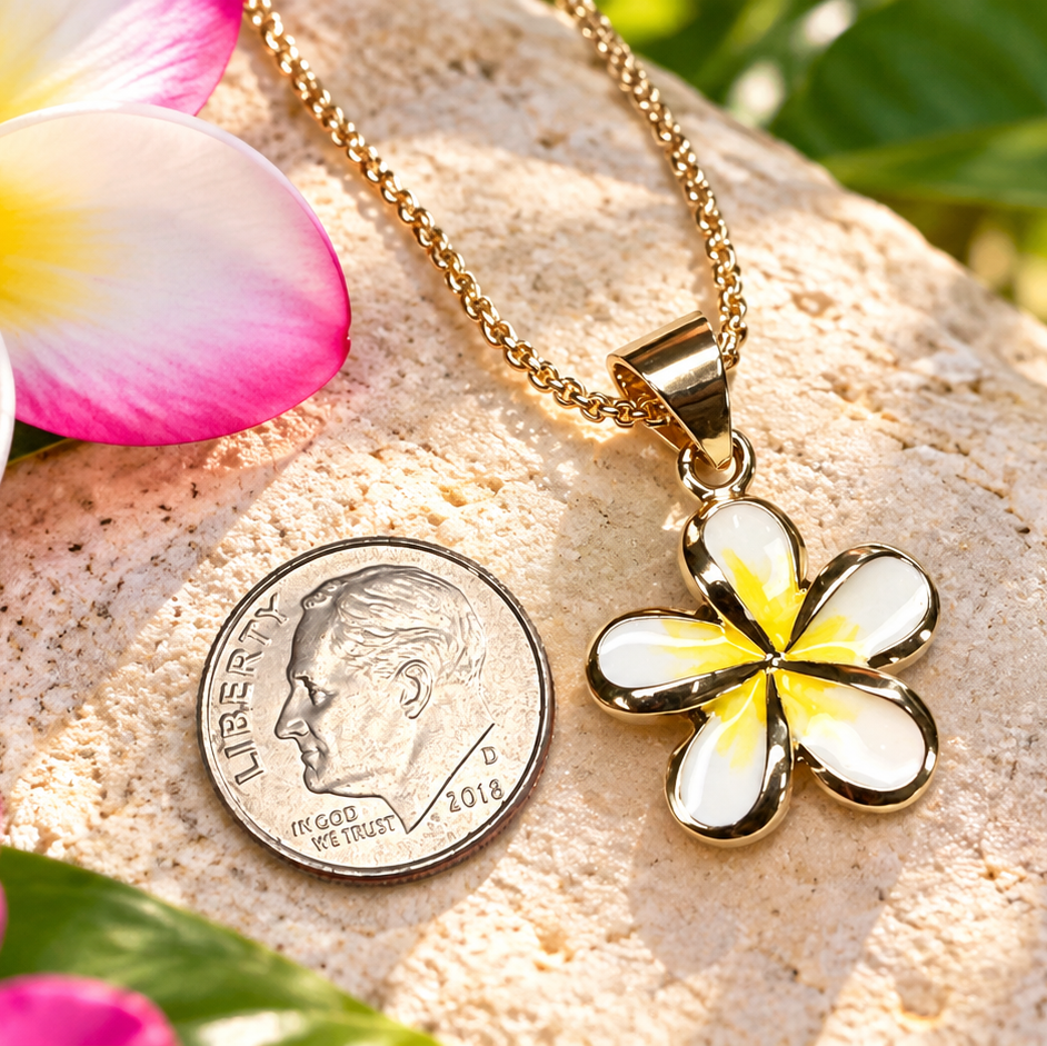 A gold-plated plumeria flower pendant necklace with white and yellow inlay, displayed next to a US dime for scale on a textured stone with tropical flowers and green leaves in the background.