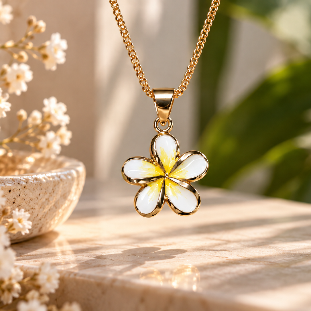 A gold-tone chain necklace with a five-petal flower pendant featuring white petals with a yellow center, displayed on a sunlit stone surface with dried flowers and green leaves in the background.