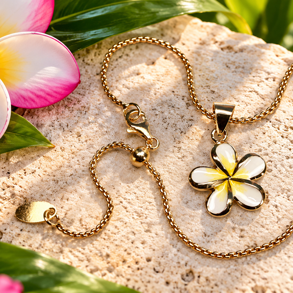 A gold-tone chain necklace with a flower-shaped pendant featuring white enamel and a yellow center, styled on a textured beige stone with tropical pink and white plumeria flowers and green leaves.
