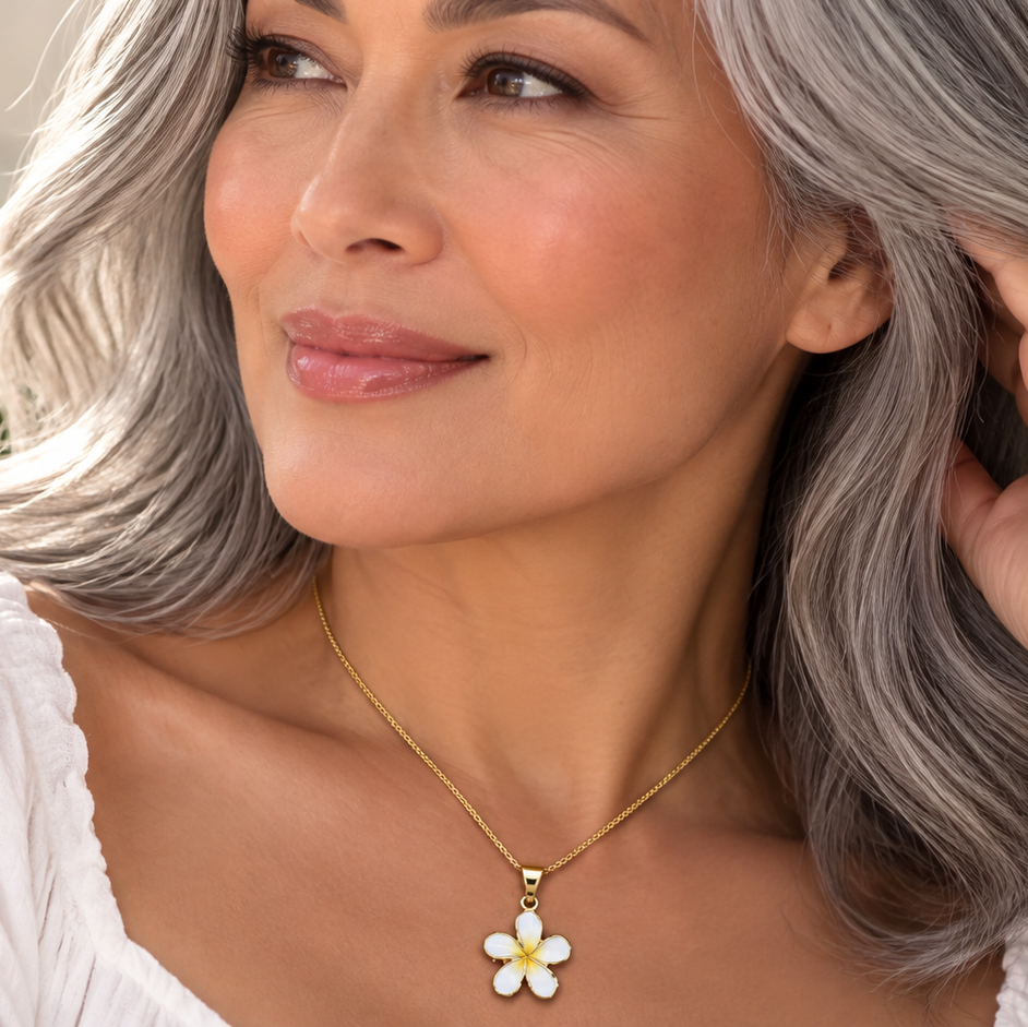 A close-up lifestyle shot of a woman with silver-gray hair wearing a delicate gold chain necklace with a white and yellow daisy flower pendant. She is wearing a white top, with a soft-focus background of green leaves and white flowers.