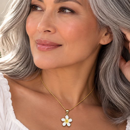 A close-up lifestyle shot of a woman with silver-gray hair wearing a delicate gold chain necklace with a white and yellow daisy flower pendant. She is wearing a white top, with a soft-focus background of green leaves and white flowers.