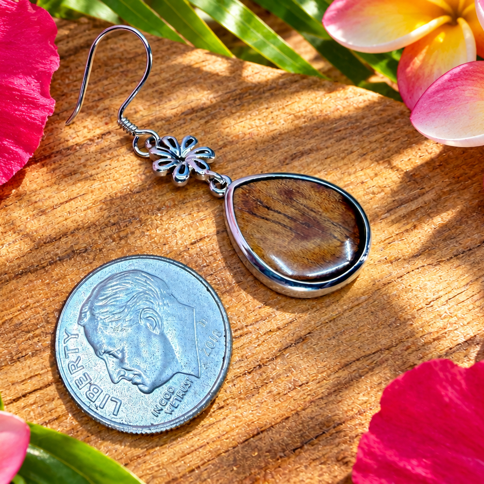 A silver dangle earring with a teardrop-shaped clear glass or acrylic pendant, displayed on a wooden surface next to a US dime for scale, surrounded by tropical hibiscus and plumeria flowers.