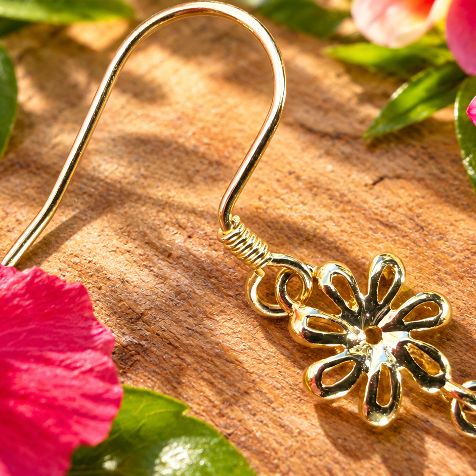 A gold-toned dangle earring with a flower-shaped charm and chain links, photographed on a wooden surface surrounded by pink hibiscus and plumeria flowers.