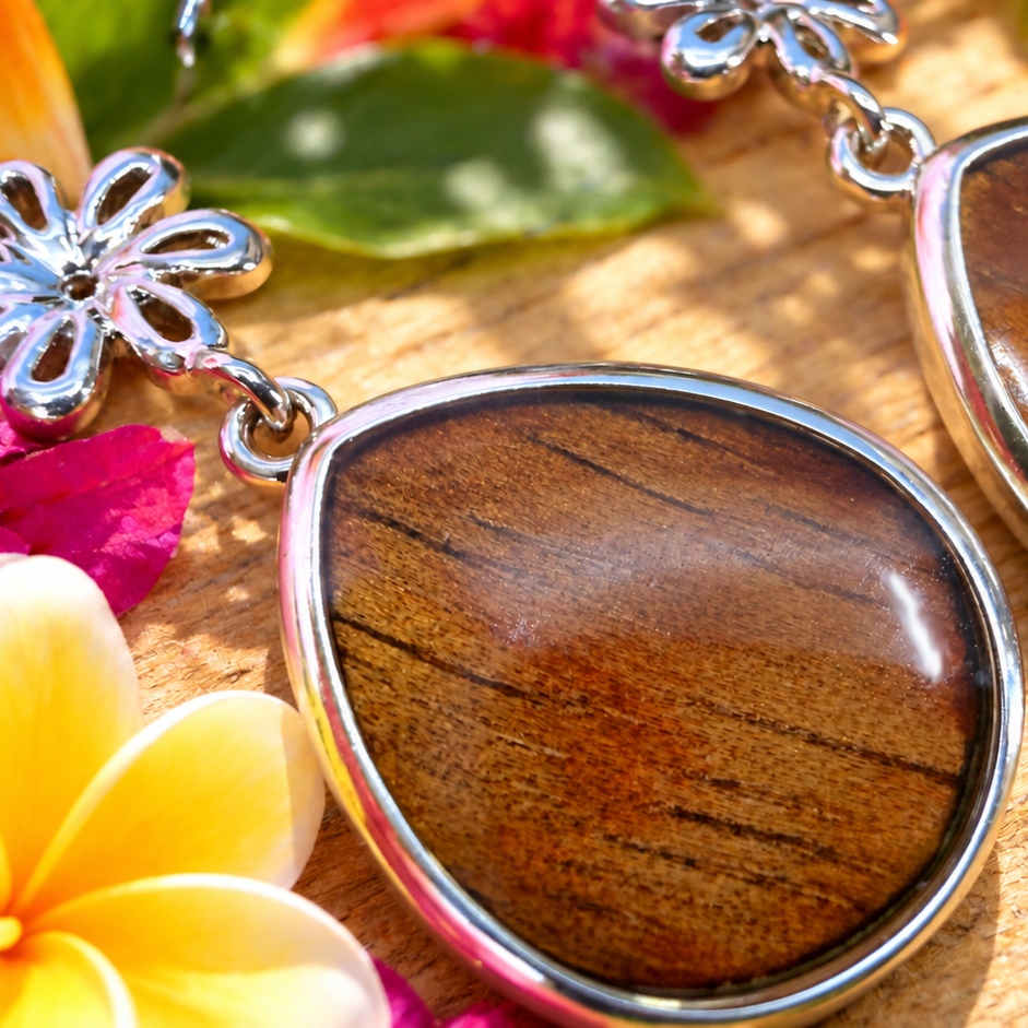A pair of teardrop-shaped dangle earrings with polished brown wood-grain cabochons set in silver-tone metal, displayed on a wooden surface surrounded by tropical flowers and leaves.