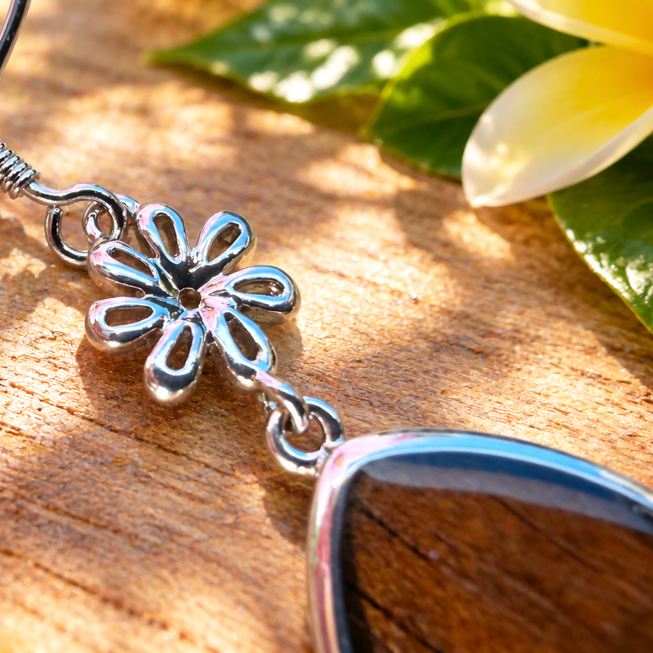 A close-up of a silver-toned dangle earring with a flower charm and a dark brown wooden bead, resting on a wooden surface with tropical flowers in the background.