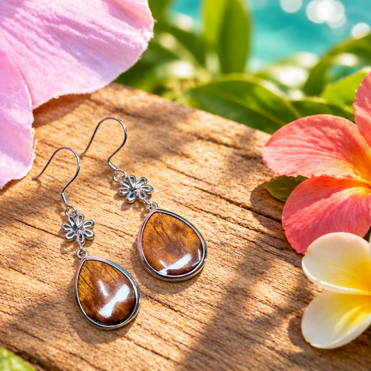 A pair of silver dangle earrings with teardrop-shaped brown tiger's eye stones, displayed on a wooden surface surrounded by tropical flowers and green leaves.