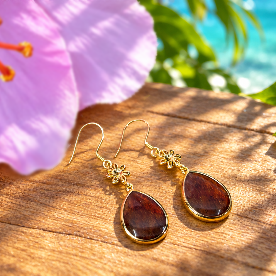 A pair of gold-tone dangle earrings with teardrop-shaped dark brown stones, resting on a wooden surface surrounded by tropical flowers and a blurred ocean background.