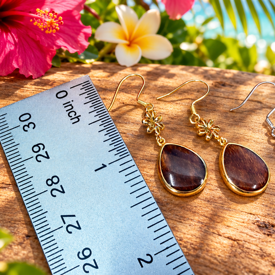 Three pairs of teardrop dangle earrings displayed on a wooden surface next to a measuring ruler, with tropical flowers and a beach scene in the background. The earrings feature brown wooden teardrop pendants in gold and silver settings.