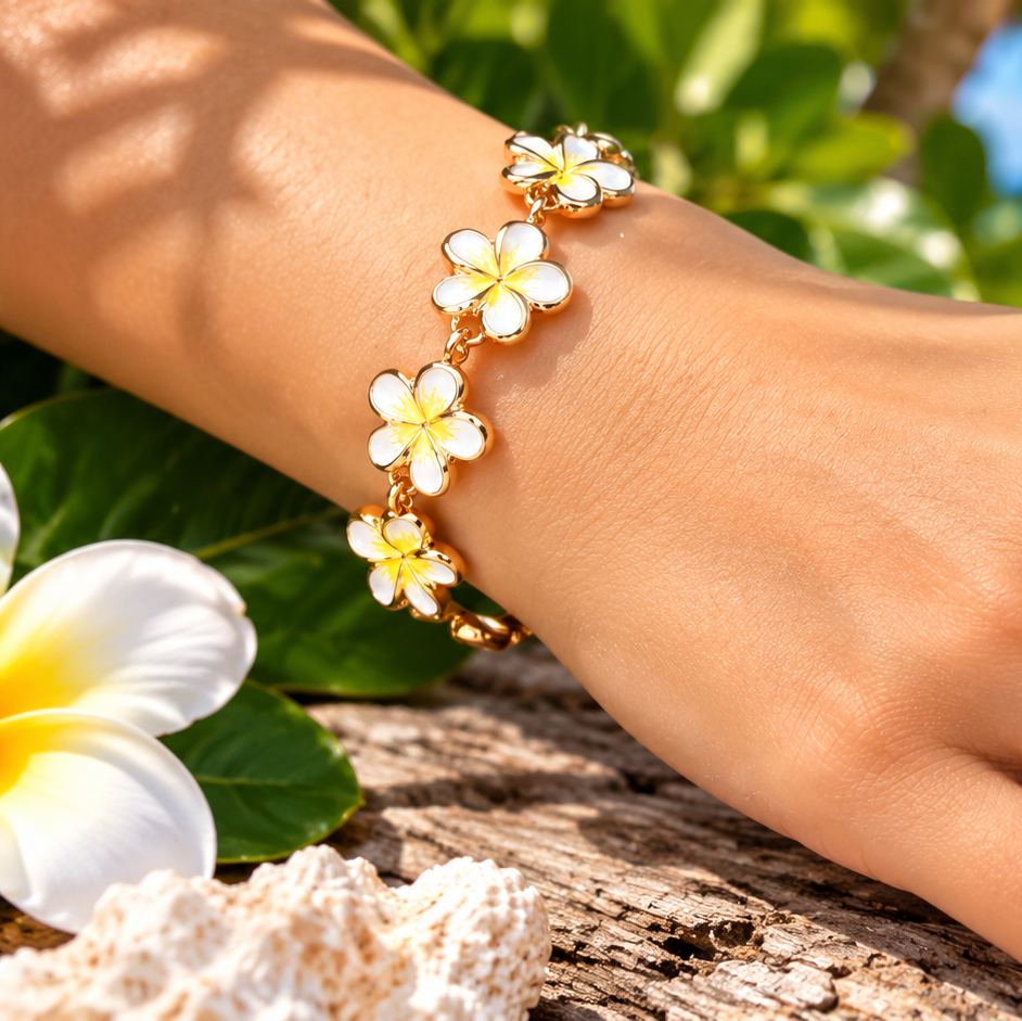 A close-up of a person's hand wearing a gold bracelet and matching flower ring, with tropical beach scenery and white plumeria flowers in the background.