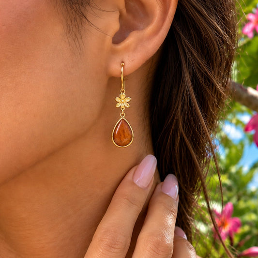 A close-up lifestyle shot of a woman wearing a gold dangle earring with a teardrop-shaped amber gemstone, set against a tropical background of pink flowers and the ocean.