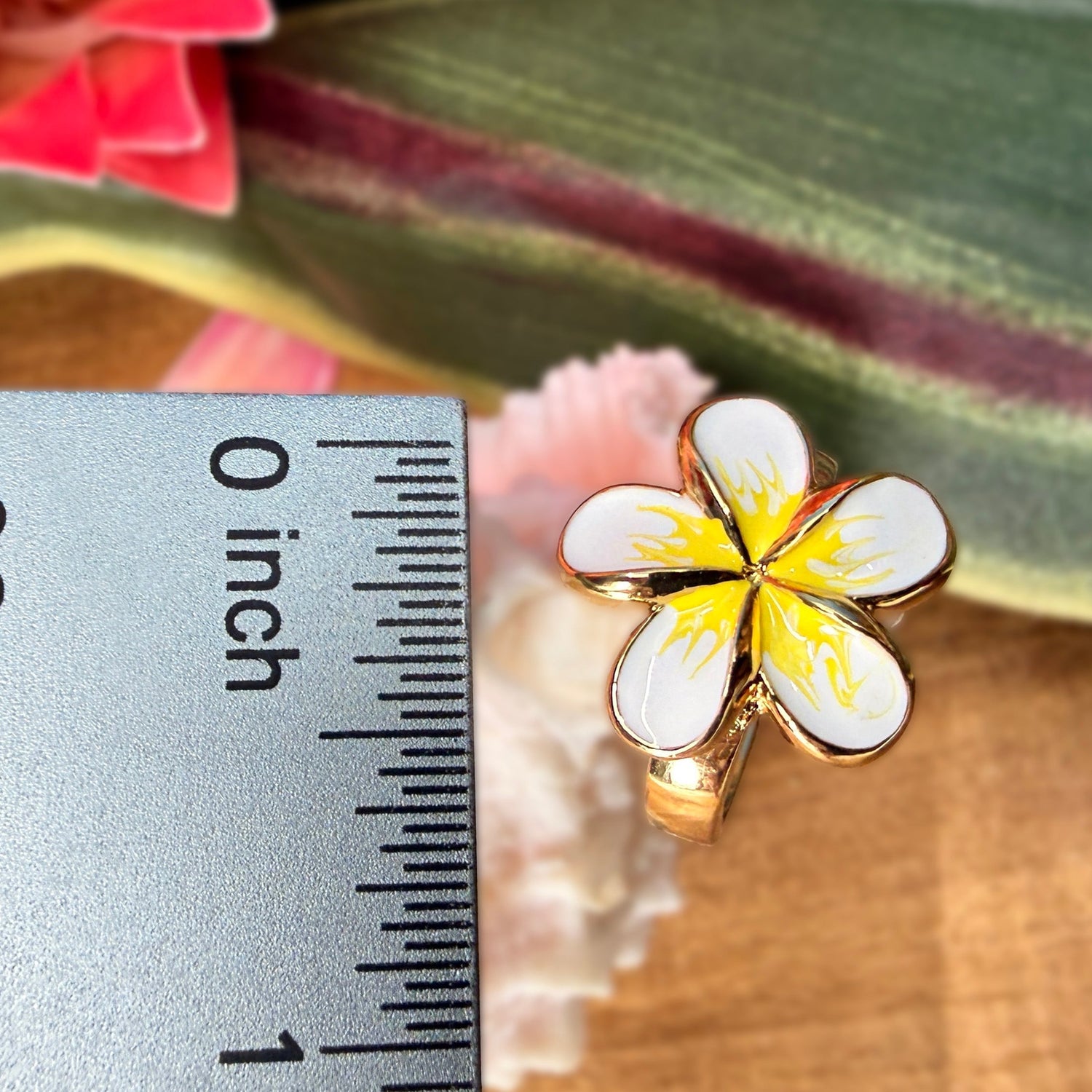 A gold-plated ring shaped like a plumeria flower with white enamel petals and a yellow center, shown next to a metal ruler for scale on a wooden surface with tropical leaves in the background.