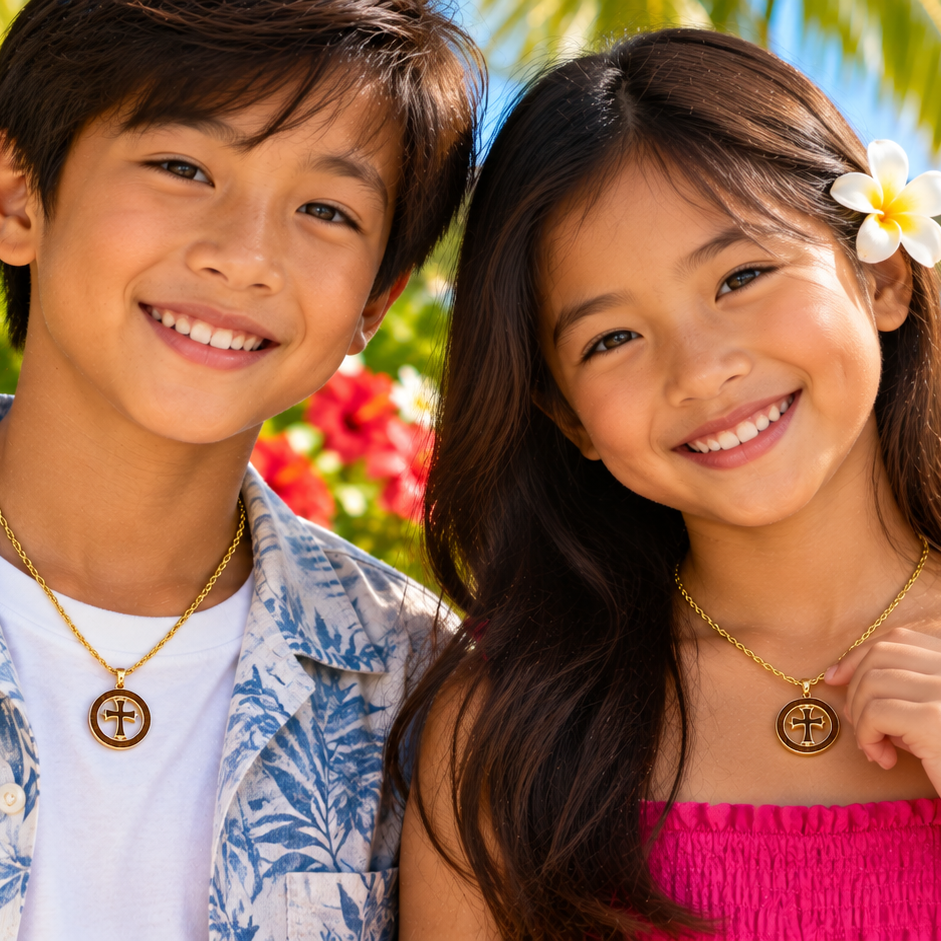 Two smiling children standing outdoors by the beach, each wearing a gold necklace with a circular cross pendant. The boy wears a white shirt with a blue tropical leaf print, and the girl wears a bright pink top.