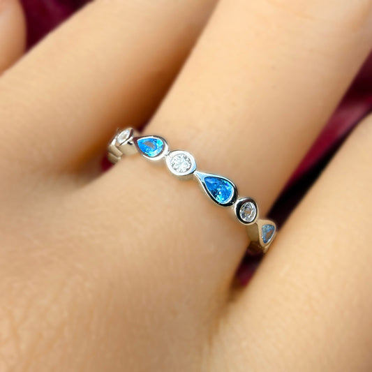 A close-up of a hand wearing a delicate silver ring adorned with small blue teardrop-shaped stones and a central clear round stone, set against a dark burgundy background.
