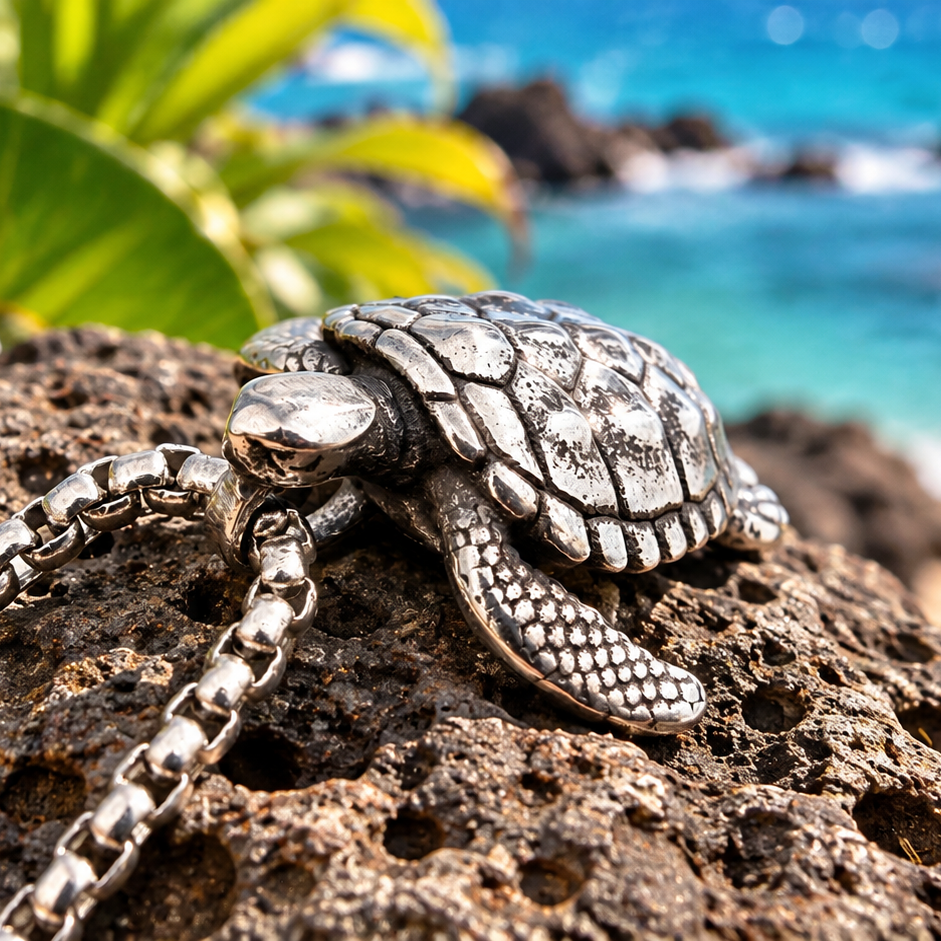 A silver sea turtle pendant necklace resting on a dark volcanic rock, with a blurred tropical beach background showing turquoise ocean water, rocks, and a white plumeria flower in the foreground.