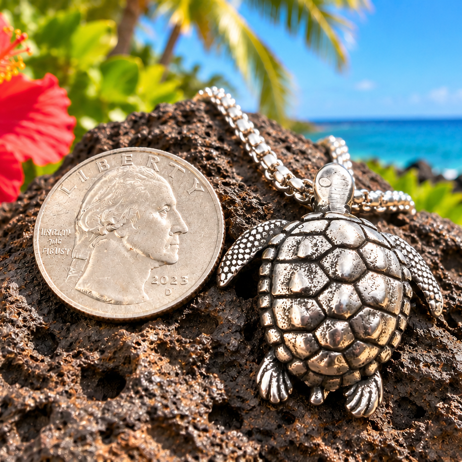A silver turtle pendant necklace is displayed on a dark volcanic rock next to a US quarter coin for scale, with a tropical beach, blue ocean, and flowers in the background.