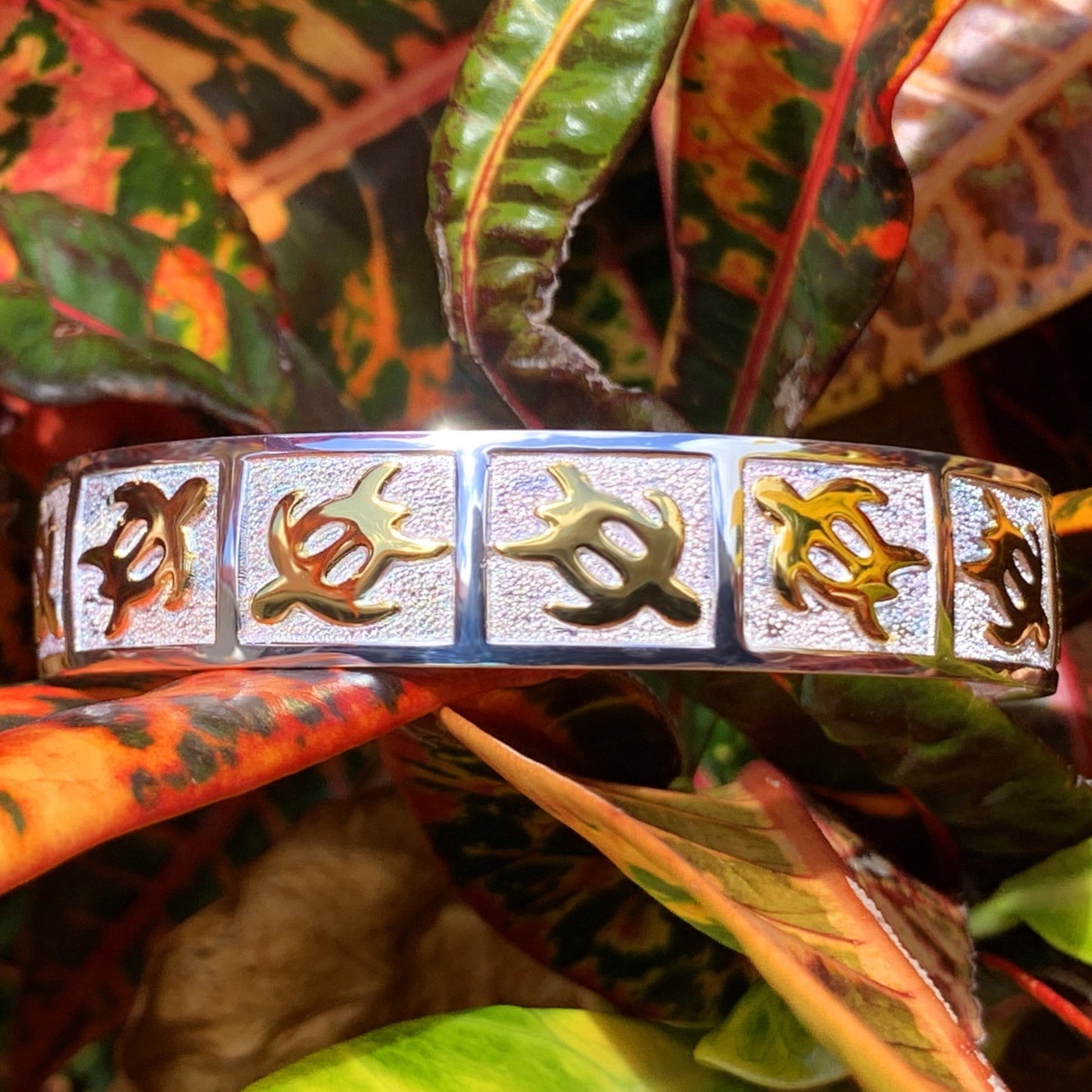 Close up of petroglyph honu cuff with gold honus in alternating directions on a pebble design background with a mirrored sterling silver base.