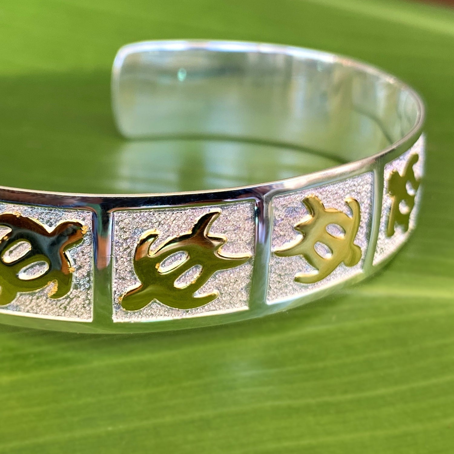 Another Close up of petroglyph honu cuff with gold honus in alternating directions on a pebble design background with a mirrored sterling silver base.